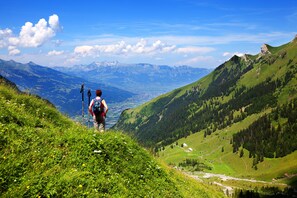 Ein Wanderer auf einem grasbewachsen Bergpfad mit Blick auf ein Tal und ferne Berge.
