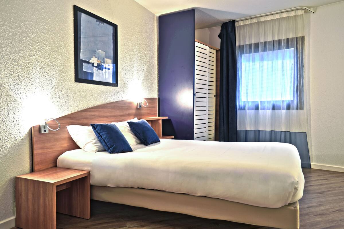 Quarto de hotel bem arrumado, com uma cama grande, mesas de cabeceira, um quadro pendurado na parede e uma janela com cortinas.
