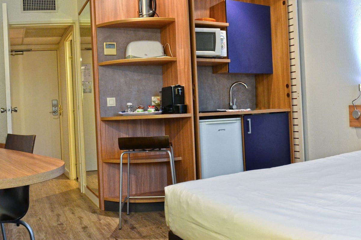 Quarto de hotel com uma cama, escrivaninha, cadeira, mini-geladeira e micro-ondas.