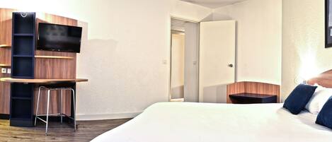 A hotel room with a bed, a TV mounted on a wooden cabinet, a small stool, and a door leading to another room.