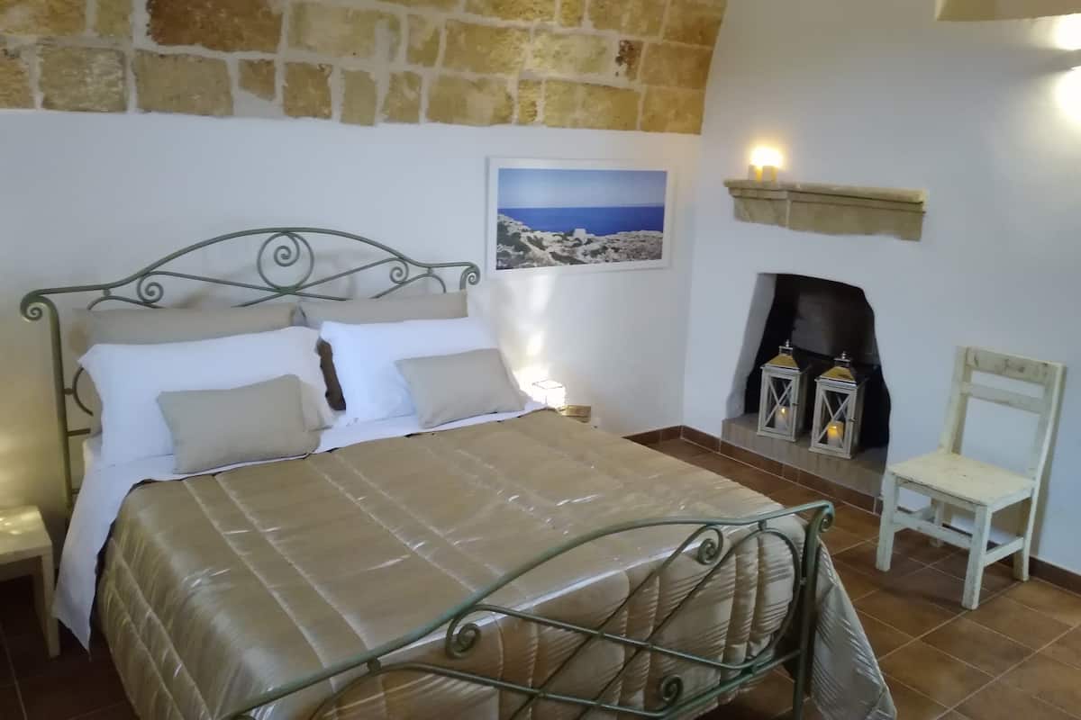 Une chambre avec un mur en pierre, un lit avec un cadre en métal, une cheminée et un tableau encadré au mur.
