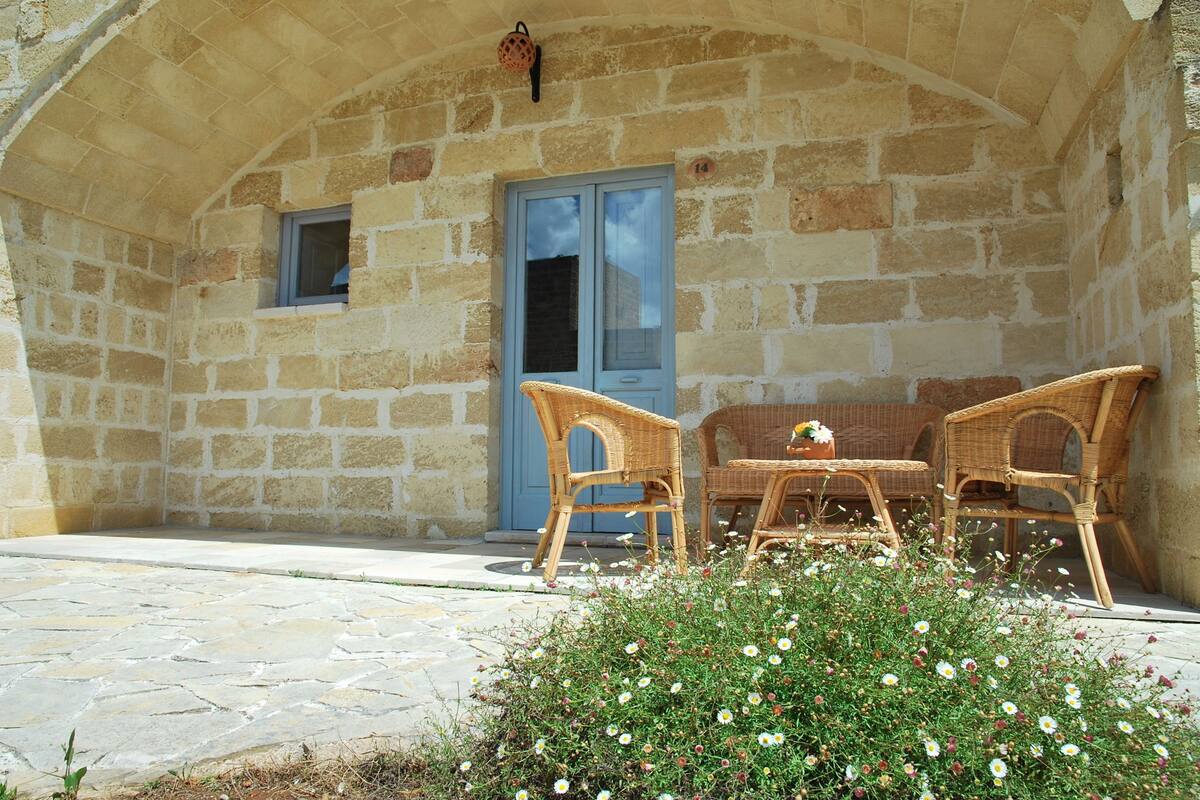 Un bâtiment en pierre avec une porte bleue et une petite fenêtre. On y trouve des chaises et une table en osier à l’extérieur.