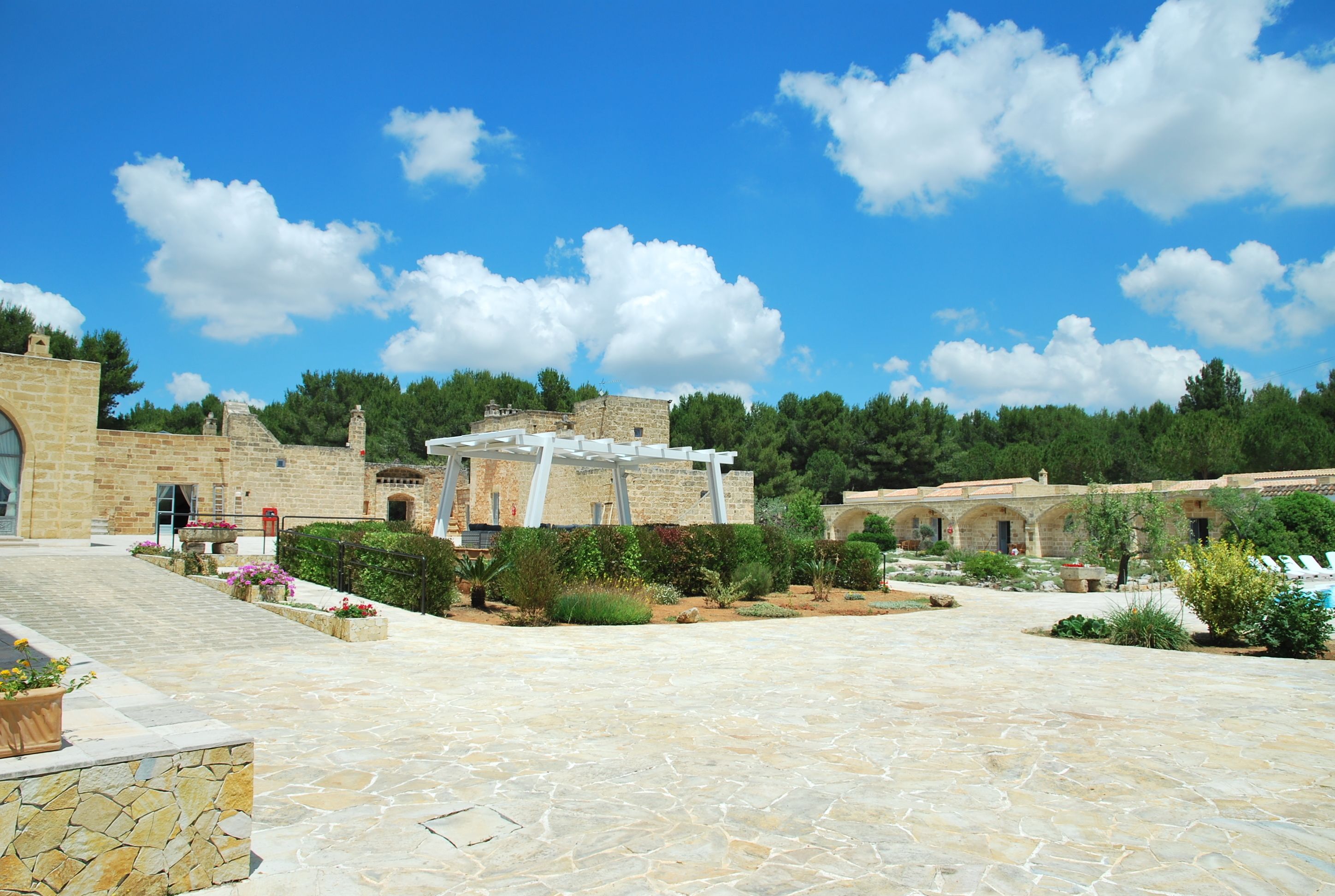 A courtyard with stone flooring, a white pergola, and stone buildings with arched doorways.