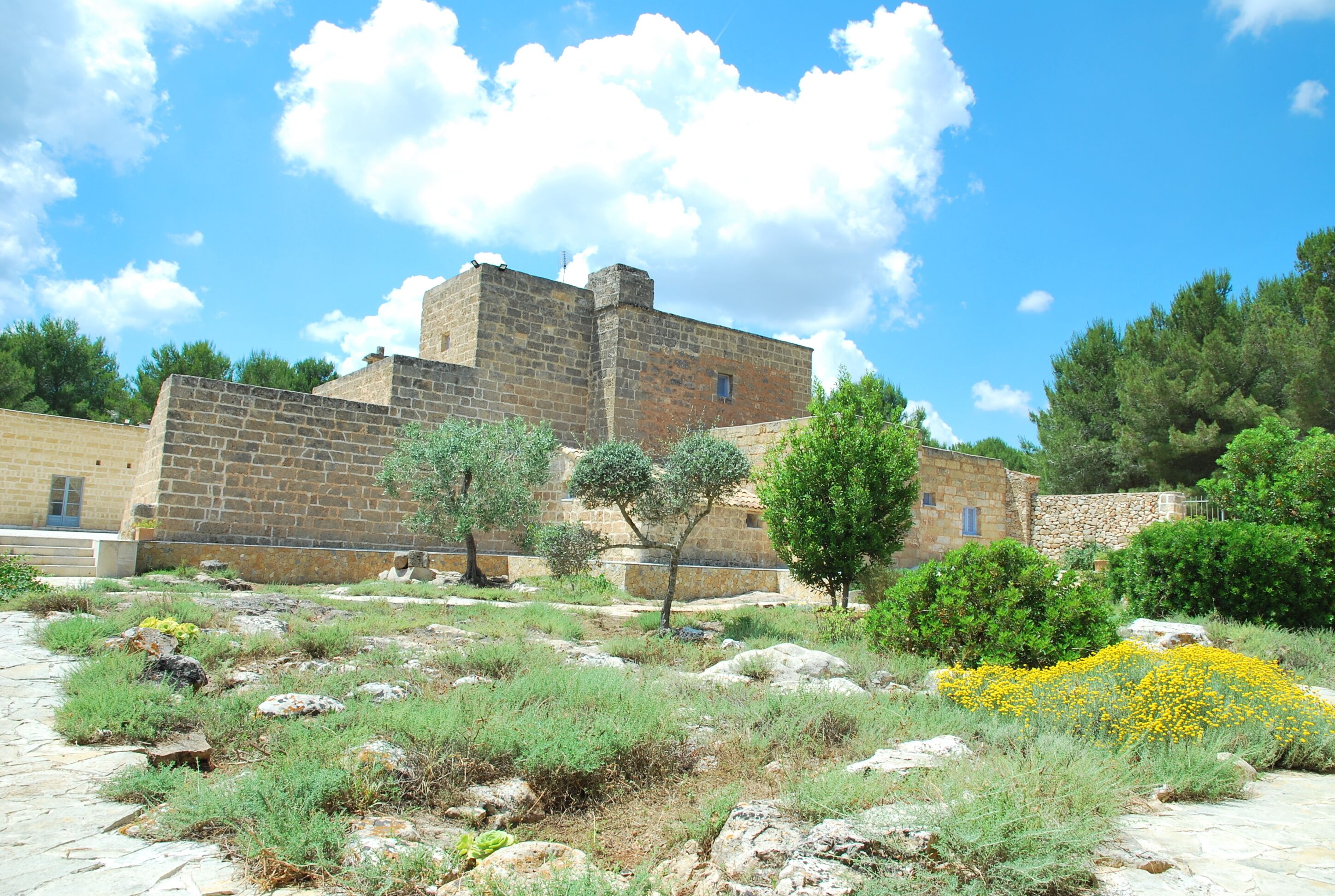 A stone building with a courtyard, surrounded by greenery and a clear sky.