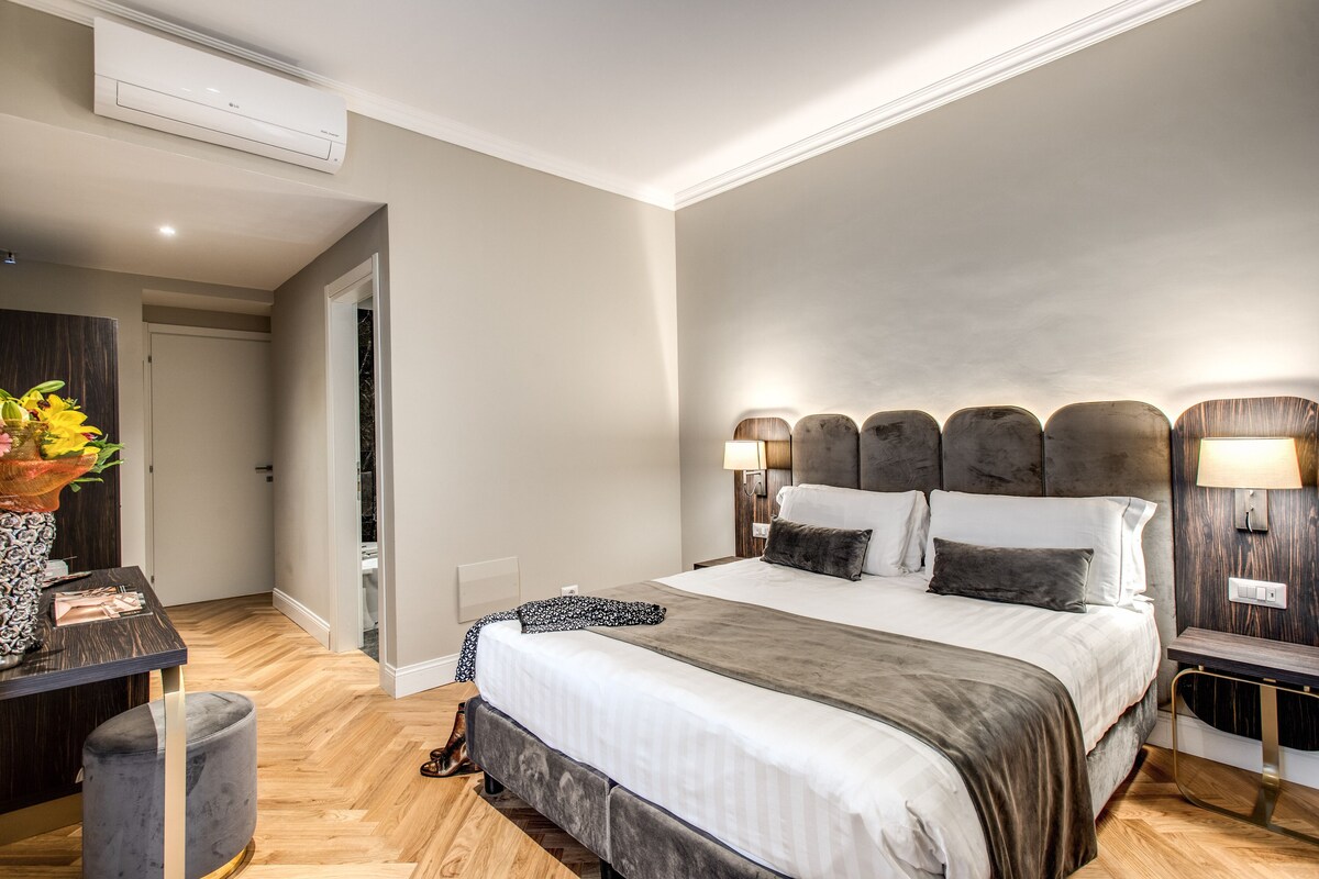 Et moderne hotelværelse med en stor seng, sengeborde med lamper, et skrivebord med en vase blomster og trægulv.
