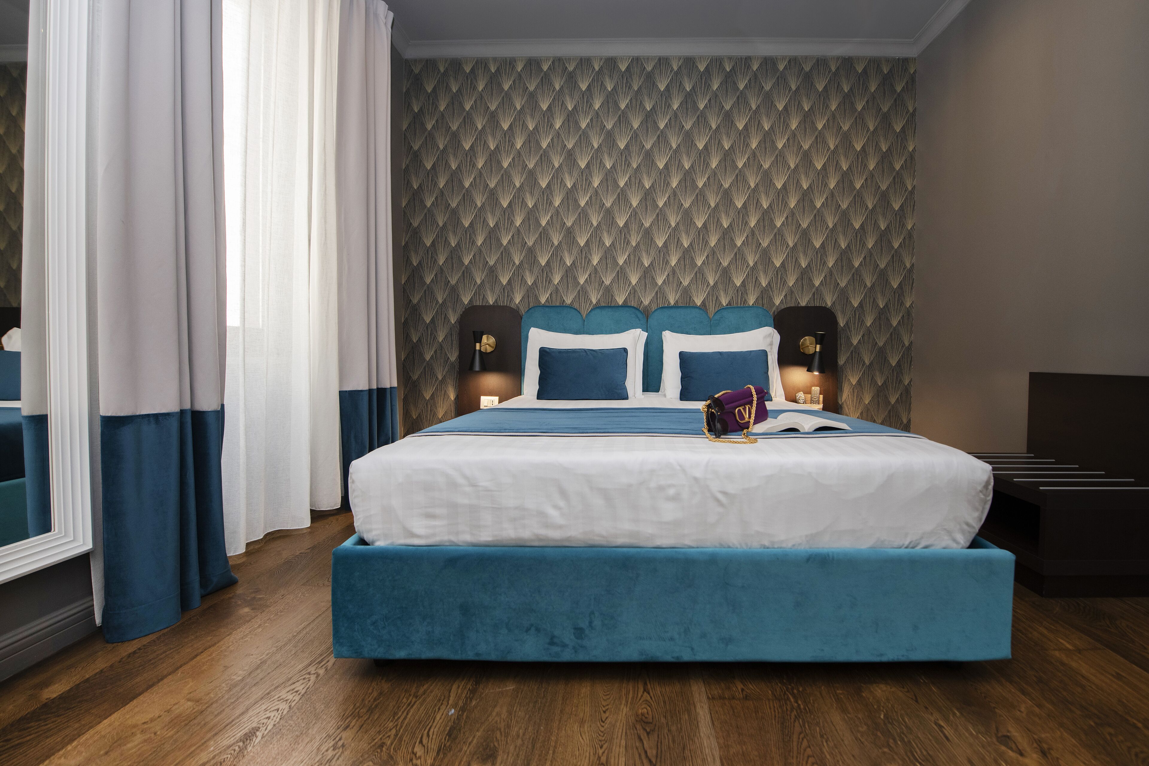 A bedroom with a large bed, two bedside tables, a nightstand, a window with curtains, and a patterned wallpaper.