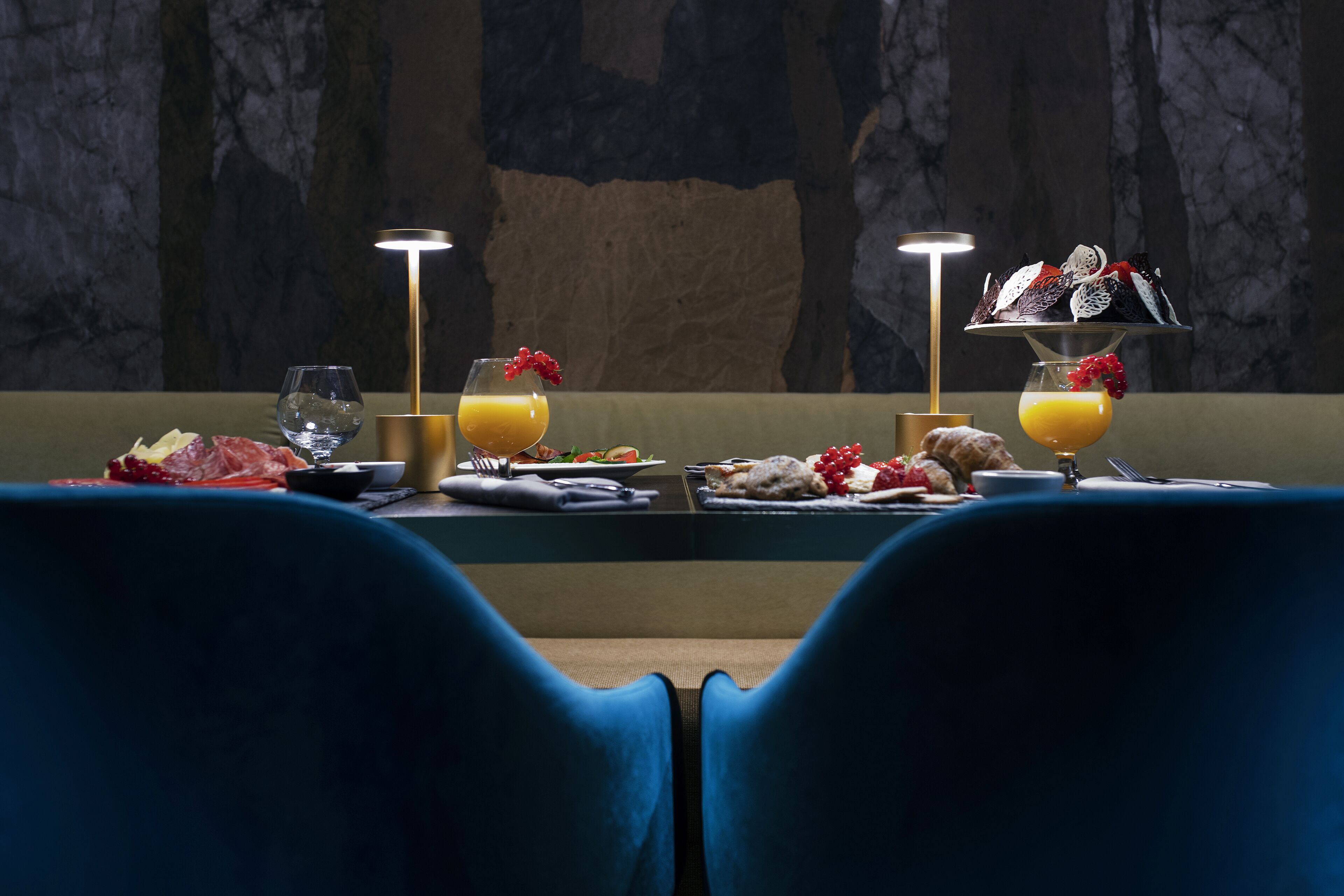 Une table à manger dressée avec des assiettes, des verres et un plateau de fruits, avec deux lampes et un mur en marbre en arrière-plan.