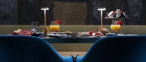 Une table à manger dressée avec des assiettes, des verres et un plateau de fruits, avec deux lampes et un mur en marbre en arrière-plan.