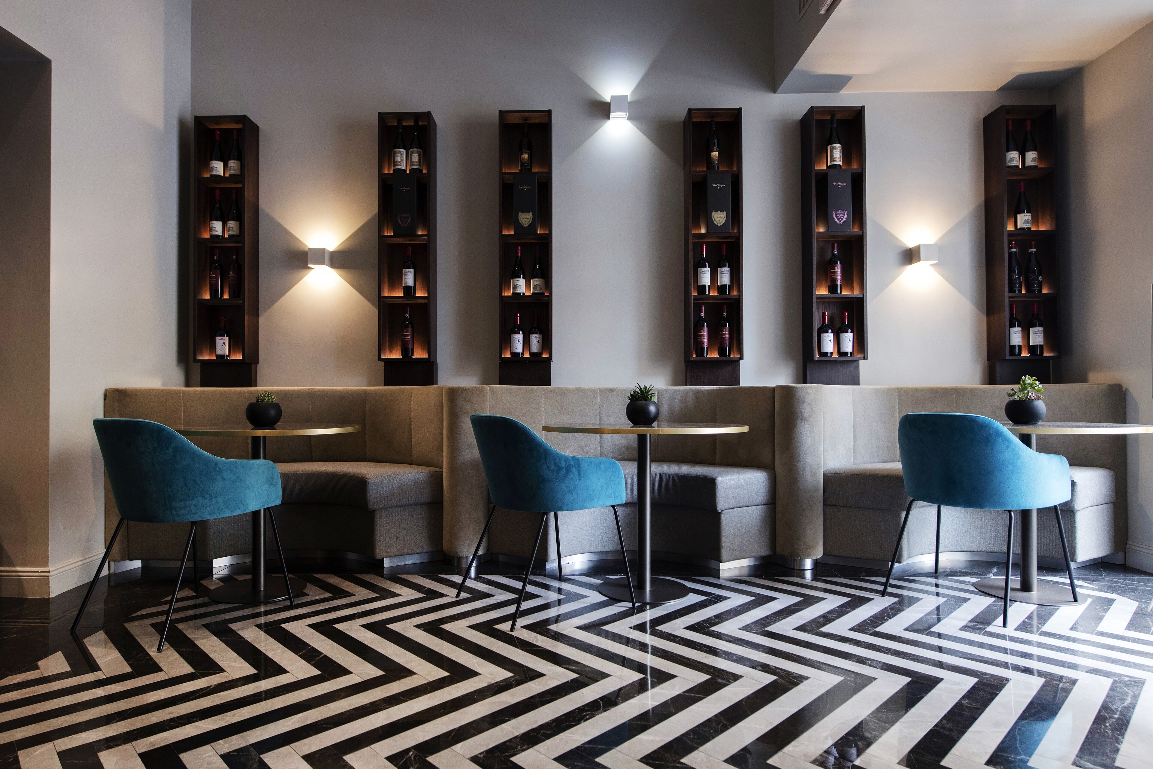 A modern lounge area with a black and white patterned floor, blue chairs, and a wine display with illuminated labels.