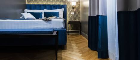 A hotel room with a large bed, a wooden floor, a patterned wall, a nightstand, and a wall-mounted lamp.