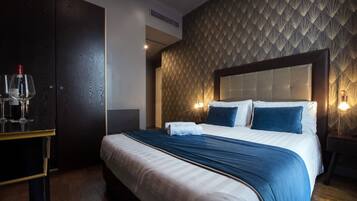 A neatly made bed with a blue bedspread, two pillows, and a headboard. There is a nightstand with a lamp and a small table with glasses and a bottle. The room has a patterned wall and a wooden floor.