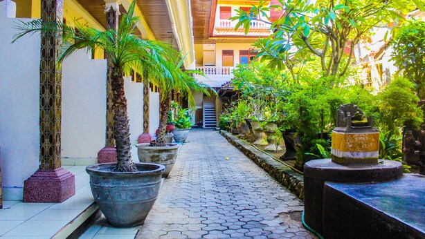 A narrow pathway flanked by potted plants and architectural columns, leading to a building with a balcony. - Bali Manik Beach Inn (Kuta)