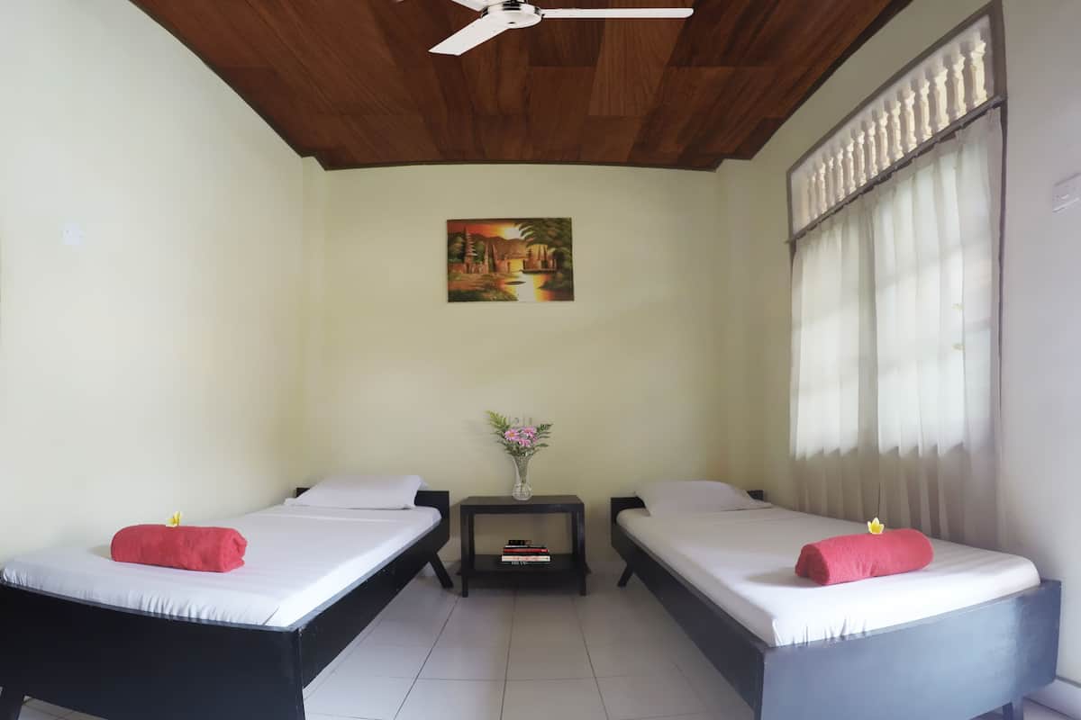 A room with two single beds, a small table with a vase of flowers, a ceiling fan, and a painting on the wall.