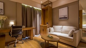 A modern hotel room with a wooden desk, a comfortable sofa, a round coffee table, and a large window with curtains.