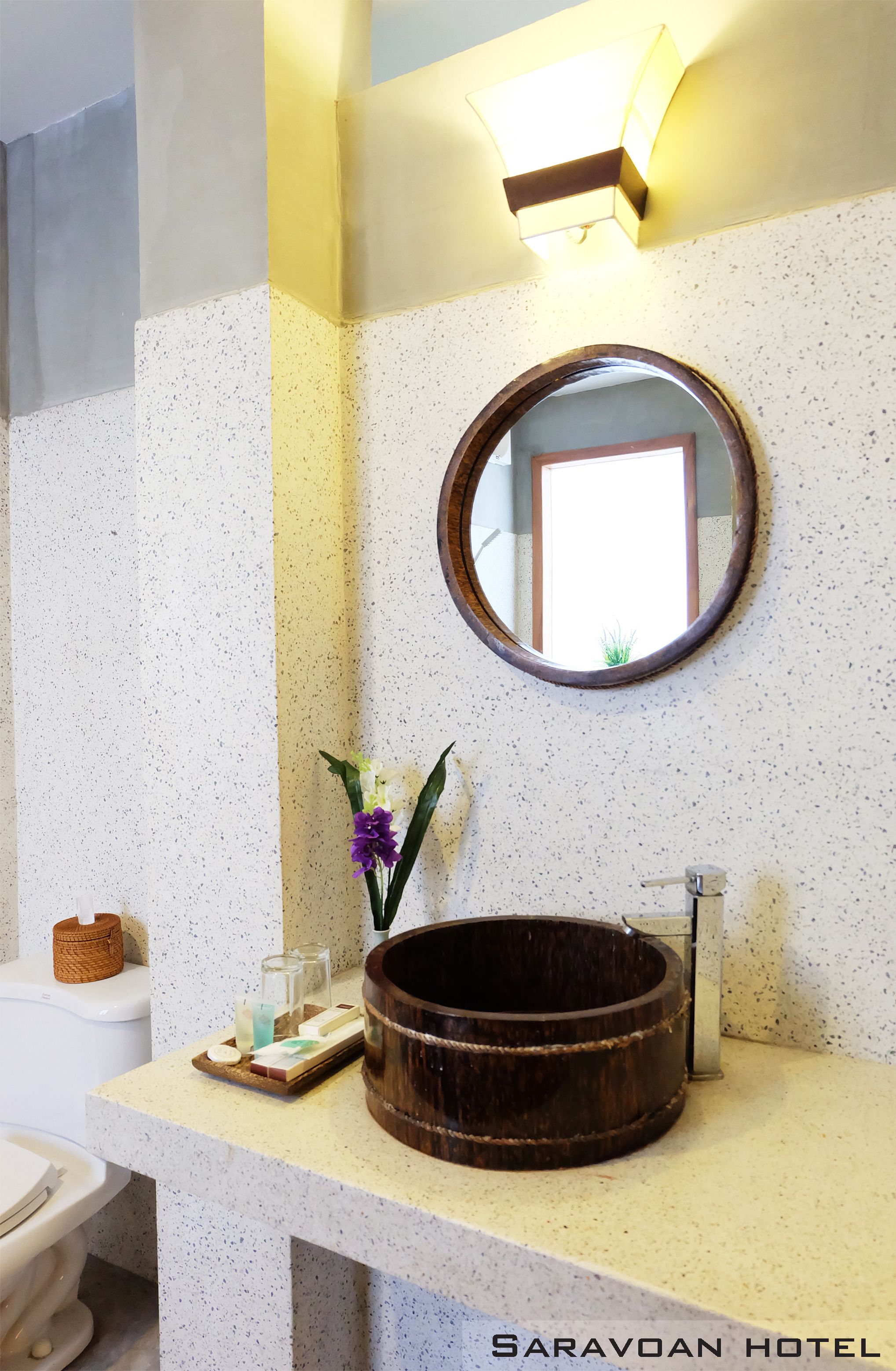 A bathroom with a round mirror, a wooden basin, and a small vase with flowers.