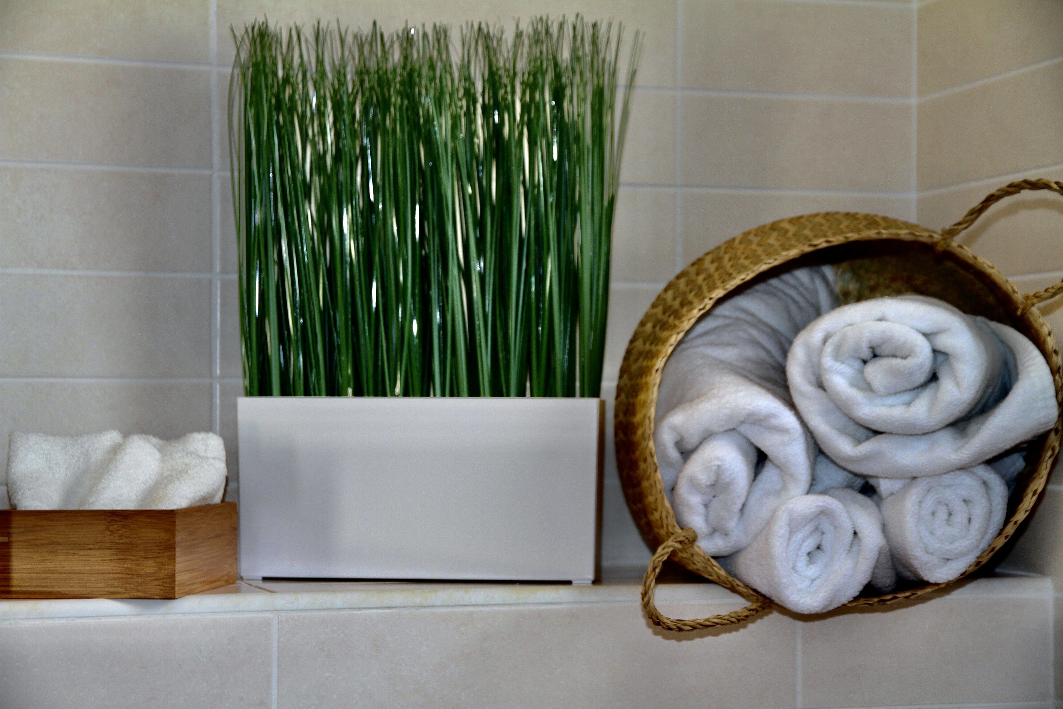 A bathroom with a white planter holding green grass, a wooden box with white towels, and a woven basket with rolled towels.