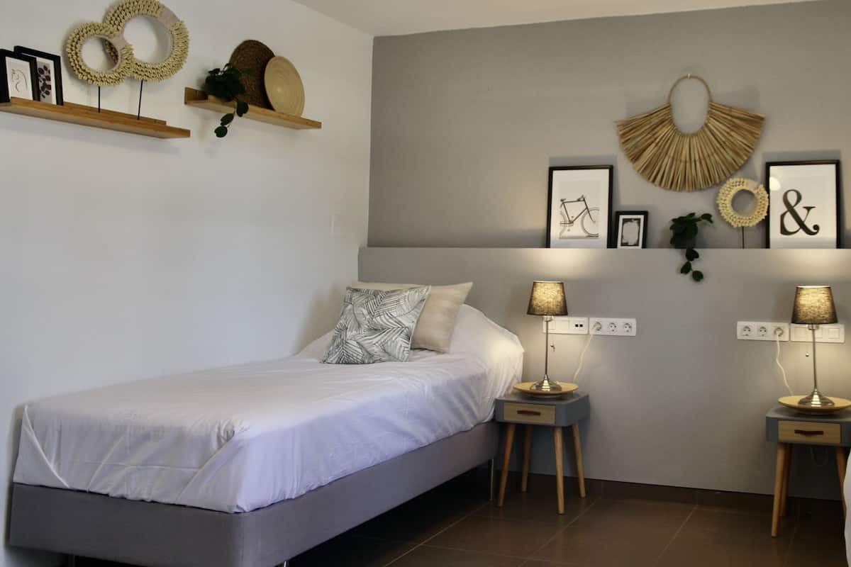 A bedroom with two single beds, bedside tables with lamps, wall-mounted shelves with decorative items, and a neutral color scheme.