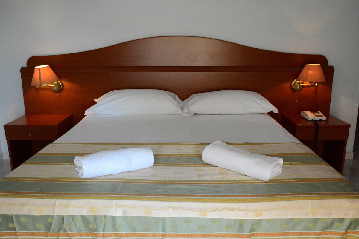 A neatly made bed with a striped bedspread, two pillows, and two rolled towels on the bed. There are two bedside tables with lamps and a telephone on one of them.