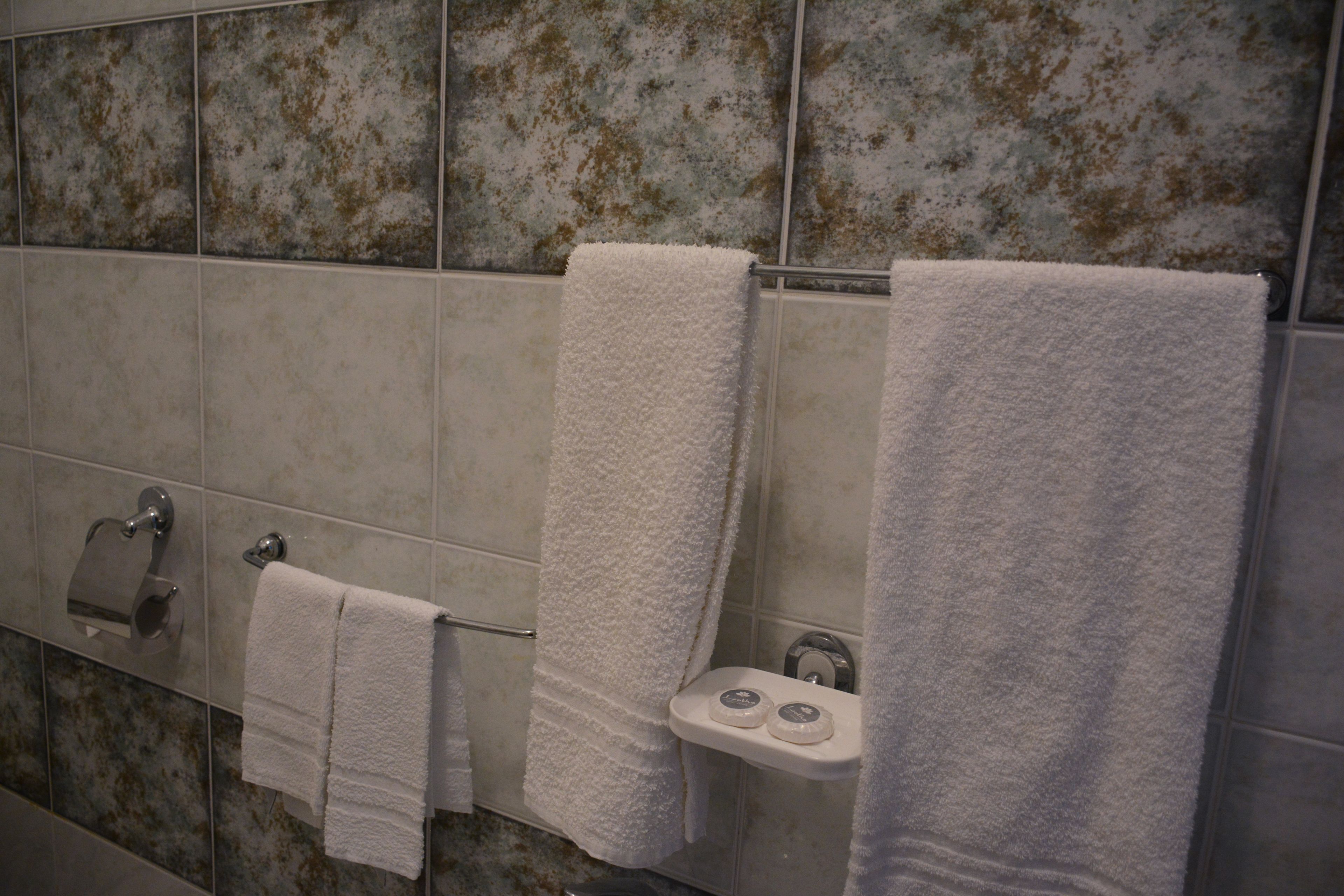 Une salle de bain avec trois serviettes blanches suspendues à un porte-serviettes, un support à papier toilette et une petite étagère sur laquelle reposent deux objets ronds.
