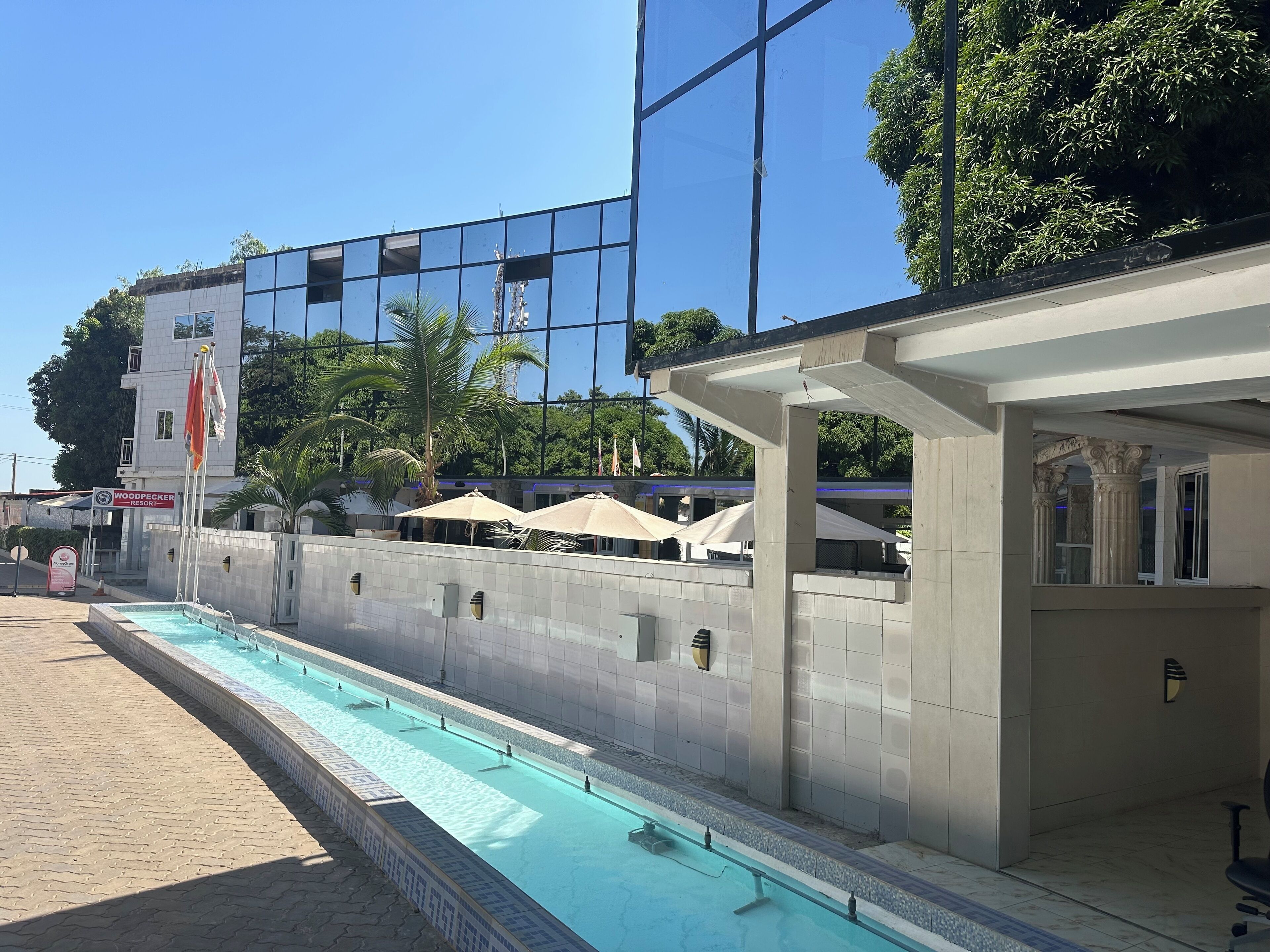 A swimming pool with a tiled deck, a building with a sign reading "Woodpecker Resort," and outdoor umbrellas.