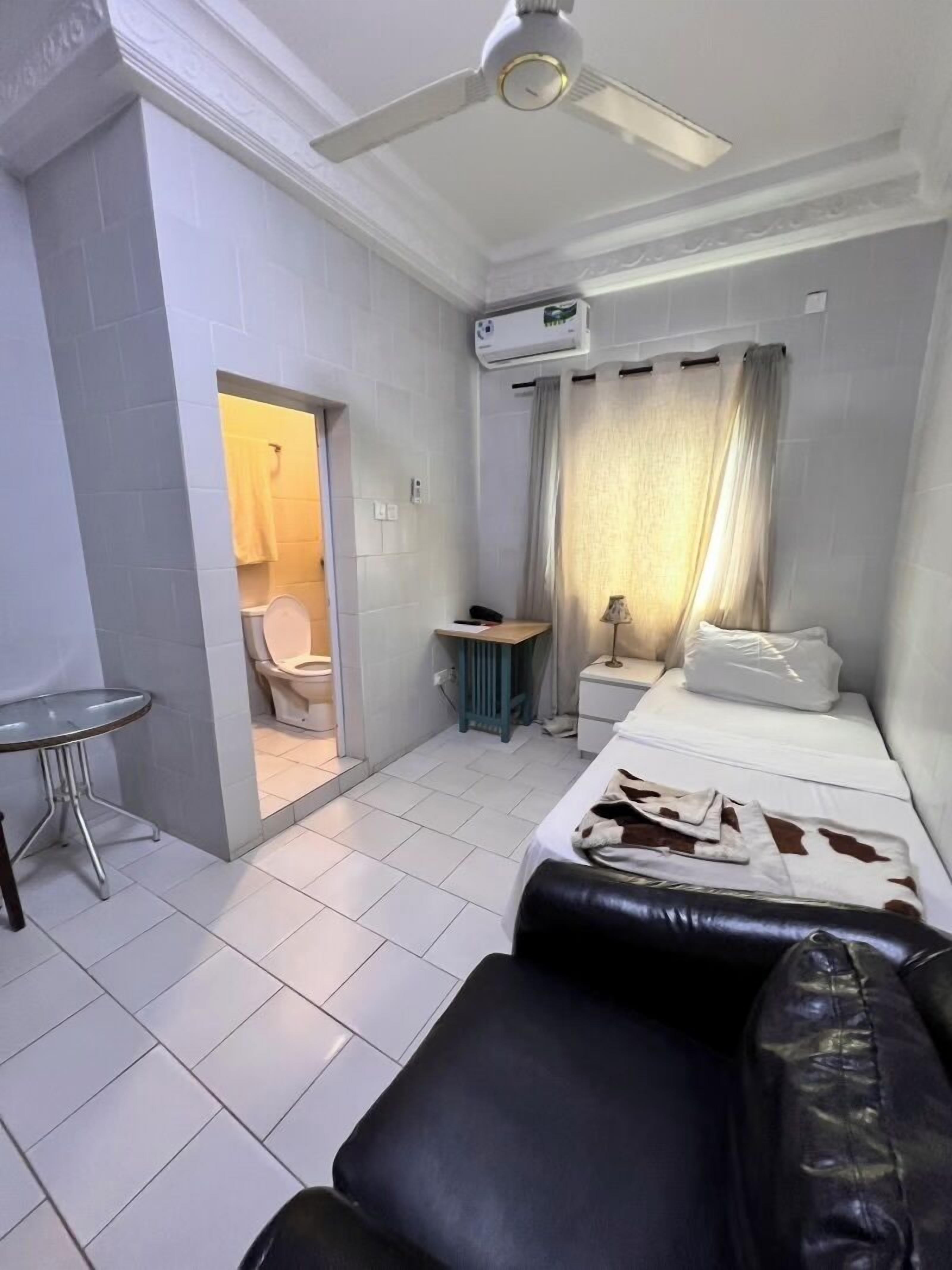 A compact hotel room with a bed, a small table, a chair, a nightstand, an air conditioning unit, and a bathroom with a toilet and shower.