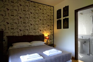 A neatly made bed with white linens, a wooden headboard, and a wall with a bird and floral wallpaper pattern. There is a bedside lamp, a framed picture, and a bathroom visible through an open door. - Villa La Ginestra (Subbiano)