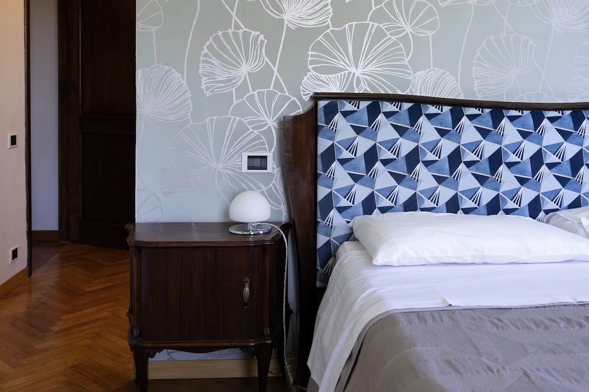 A bedroom with a patterned wall, a wooden headboard, a bedside table with a lamp, and a patterned rug.