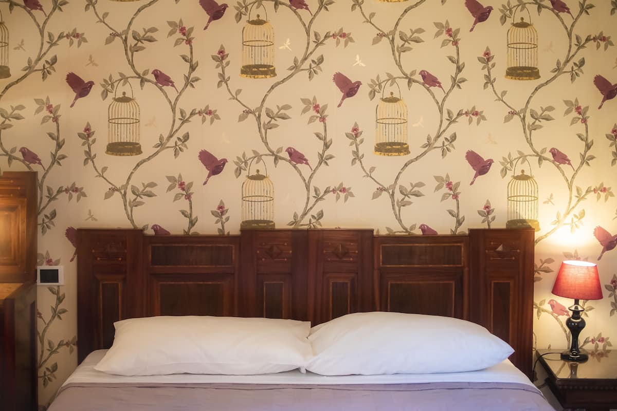 A bedroom with a floral wallpaper, a wooden headboard, a white bed with two pillows, and a nightstand with a lamp.