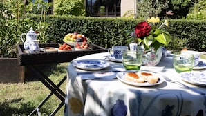 An outdoor dining area with a table set for two, surrounded by lush greenery and a building with arched windows.