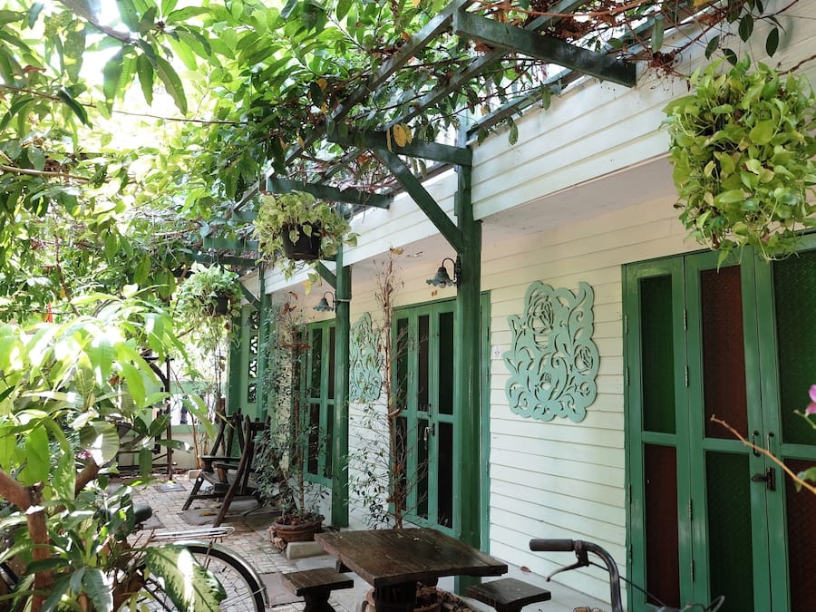 A covered outdoor area with wooden benches, hanging plants, and green doors.