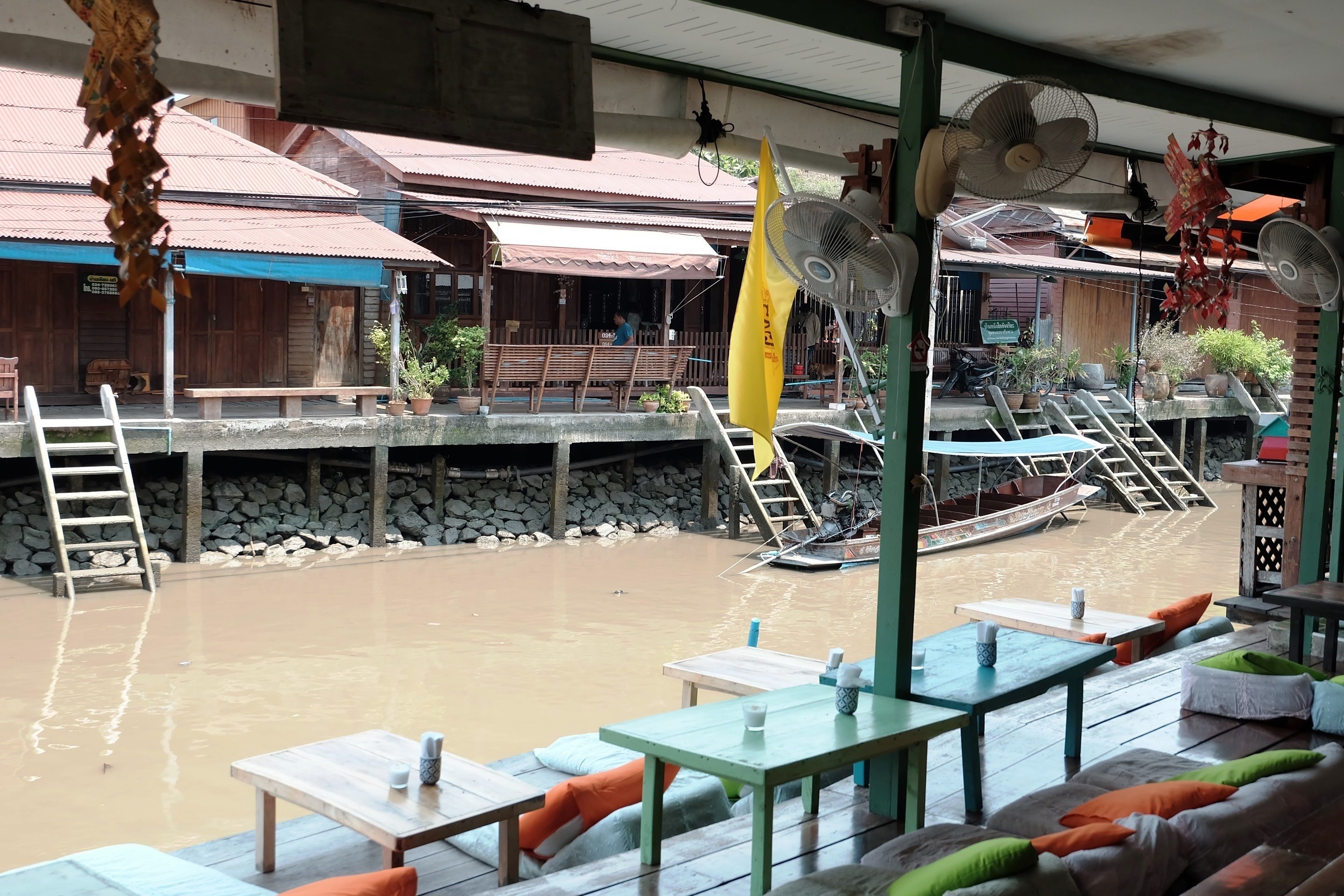 A riverside restaurant with outdoor seating, stone steps leading to a wooden deck, and a boat docked in the background.
