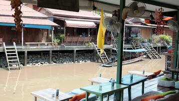 A riverside restaurant with outdoor seating, stone steps leading to a wooden deck, and a boat docked in the background.