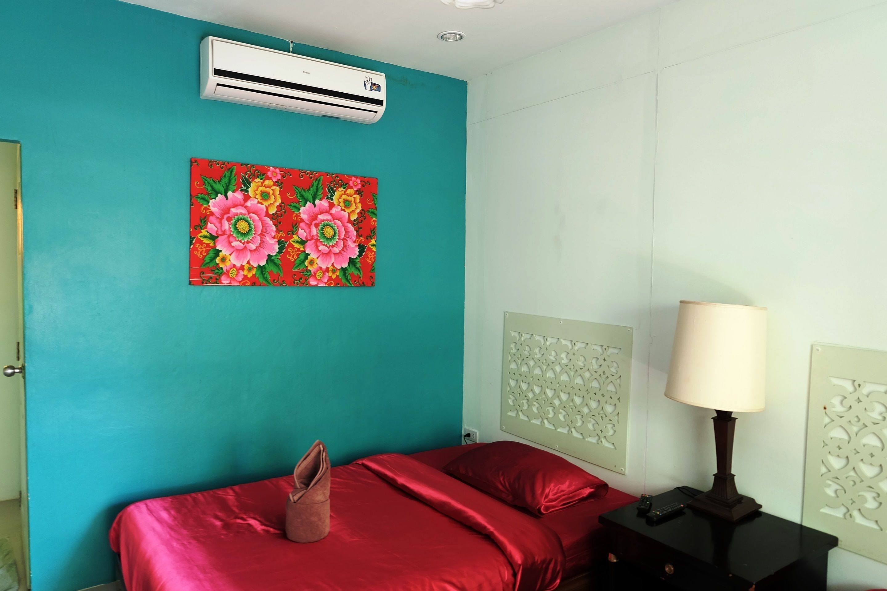 A room with teal walls, a red bed, a pillow, a lamp, an air conditioner, and a floral painting.