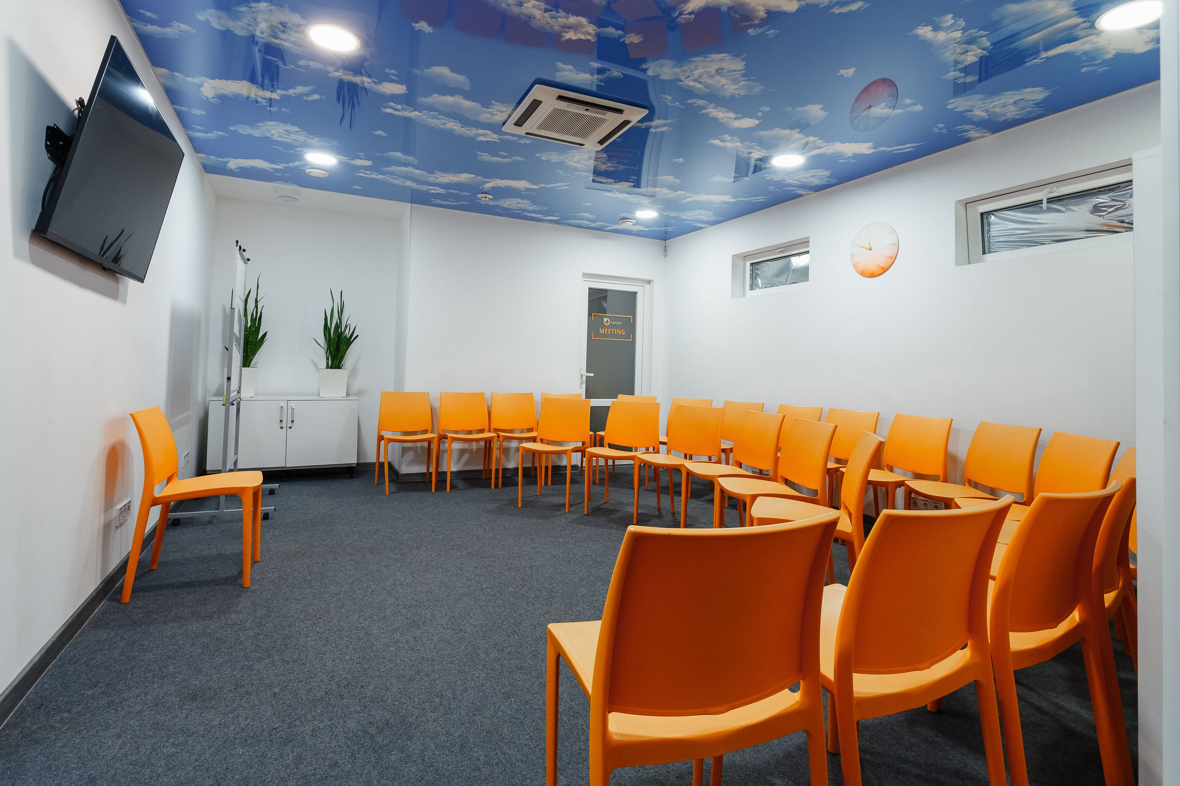 Une pièce avec une table ronde et des chaises orange, un téléviseur fixé au mur et un plafond décoré de motifs nuageux.