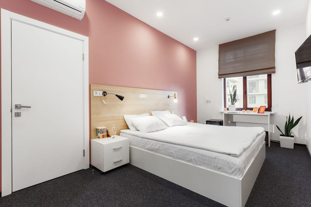 Un dormitorio moderno con una cama grande, mesitas de noche, un escritorio y una ventana con vistas a edificios.
