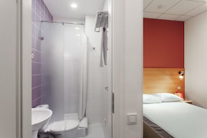 A bathroom with a shower, toilet, and sink. There is a bed with a wooden headboard and a nightstand with a lamp. - Orange Hotel (Odesa)
