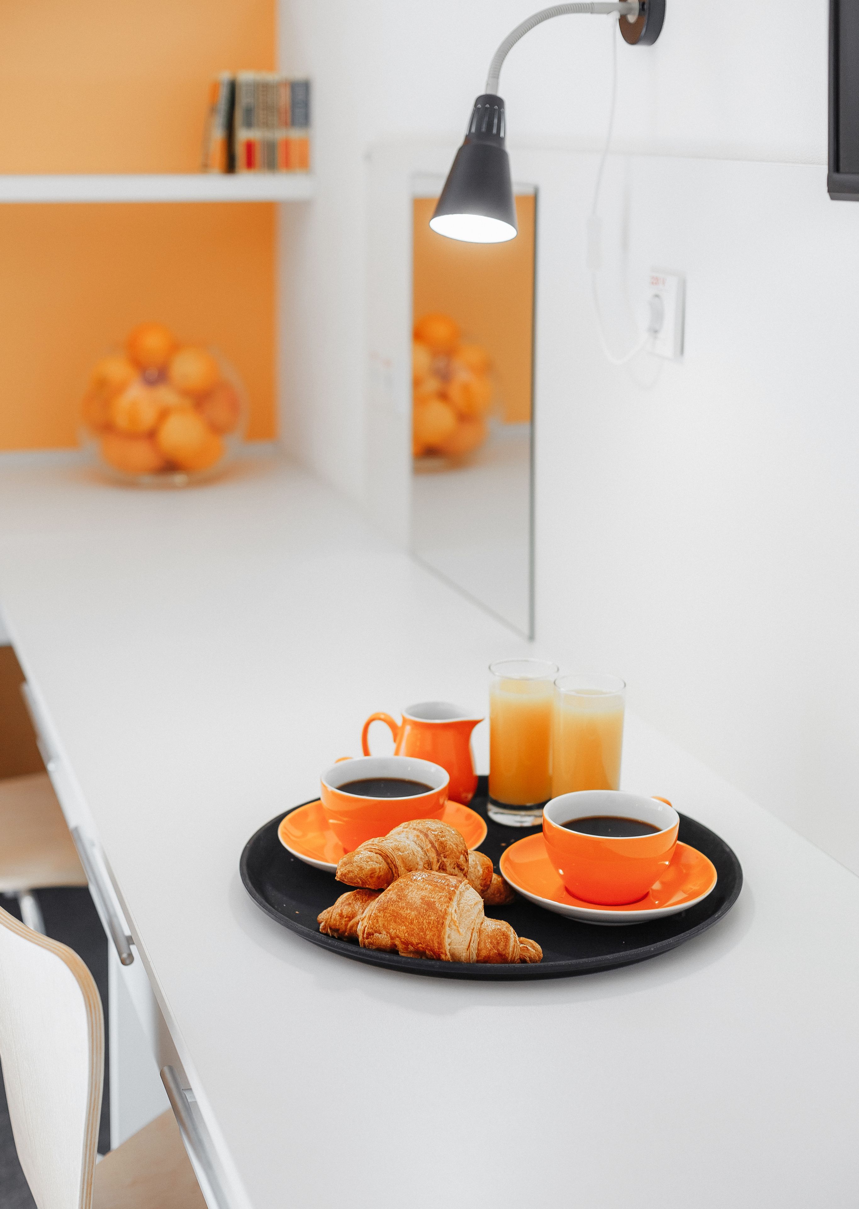 A breakfast tray with croissants, orange cups, and orange juice on a white table in a modern room with a mirror and orange decor.