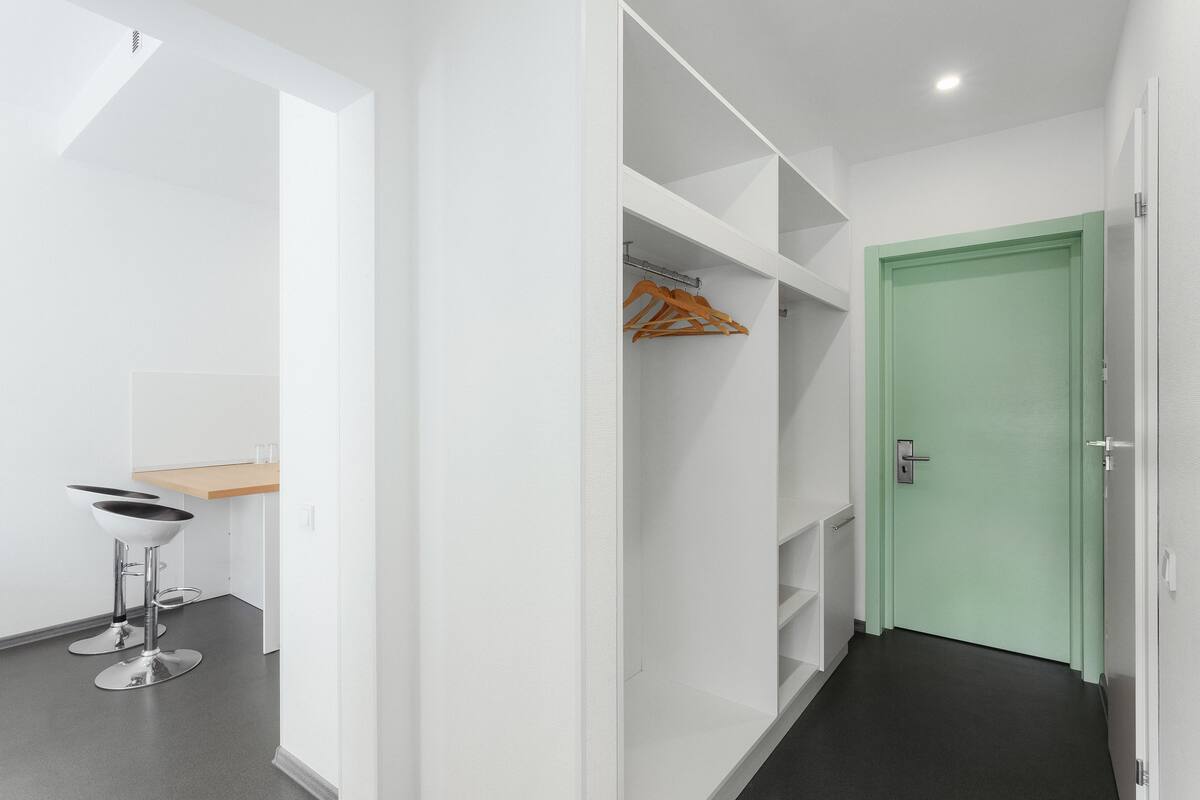 Un pasillo con una puerta verde, paredes blancas y suelo negro. Hay dos sillas de metal y un perchero de madera.