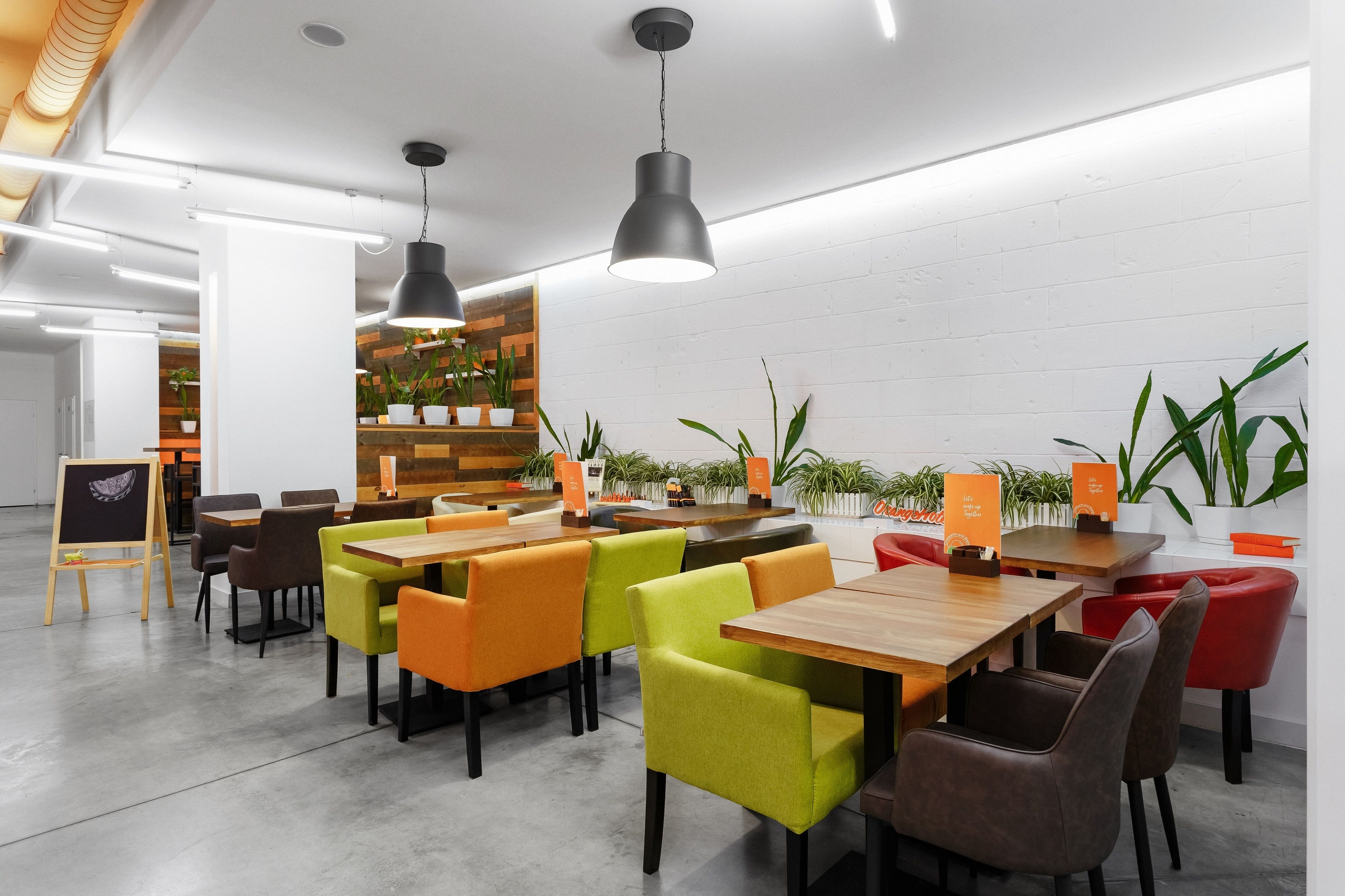 A modern cafe with wooden tables and colorful chairs, potted plants, and pendant lighting.