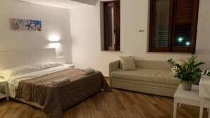 A bedroom with a bed, a sofa, a small table, and a window with shutters. - Hotel Il Pirata (Cinisi)