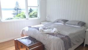 A bedroom with a bed, a nightstand, a wooden chest, and a window with a view of trees and water. - Lakeview Holiday Home (Rotorua)