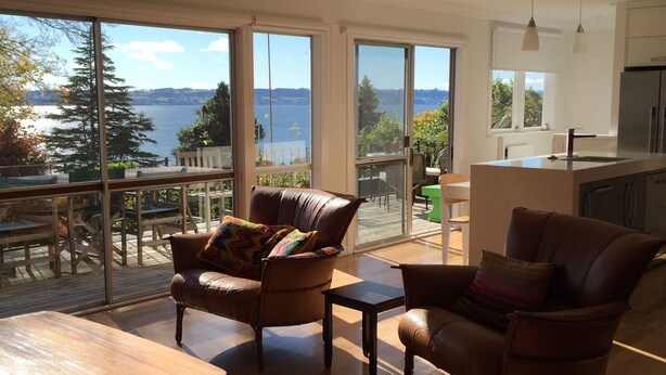 A spacious living room with large windows overlooking a lake, furnished with leather chairs and a wooden table. - Lakeview Holiday Home (Rotorua)