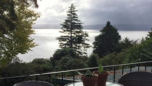 A balcony with wicker chairs and a glass table, overlooking a lake with trees and a cloudy sky. - Lakeview Holiday Home (Rotorua)