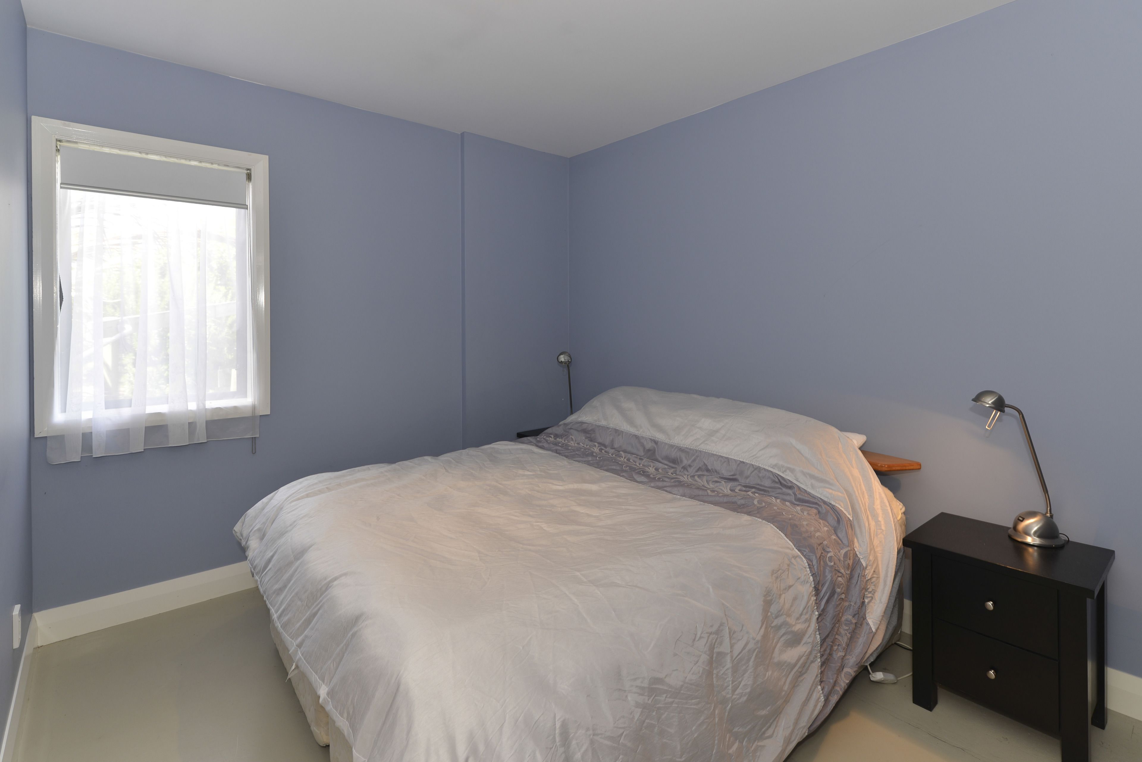 Un dormitorio con cama, mesita de noche, lámpara y ventana con cortinas translúcidas.