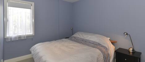Une chambre à coucher avec un lit, une table de chevet, une lampe et une fenêtre ornée de rideaux légers.
