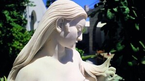 A statue of a woman with long hair, set against a backdrop of greenery and a building.