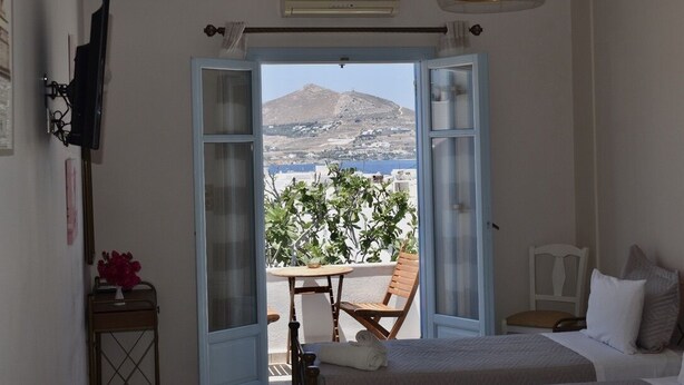 A bedroom with a bed, a chair, a table, and a view of a mountain and sea through open doors.