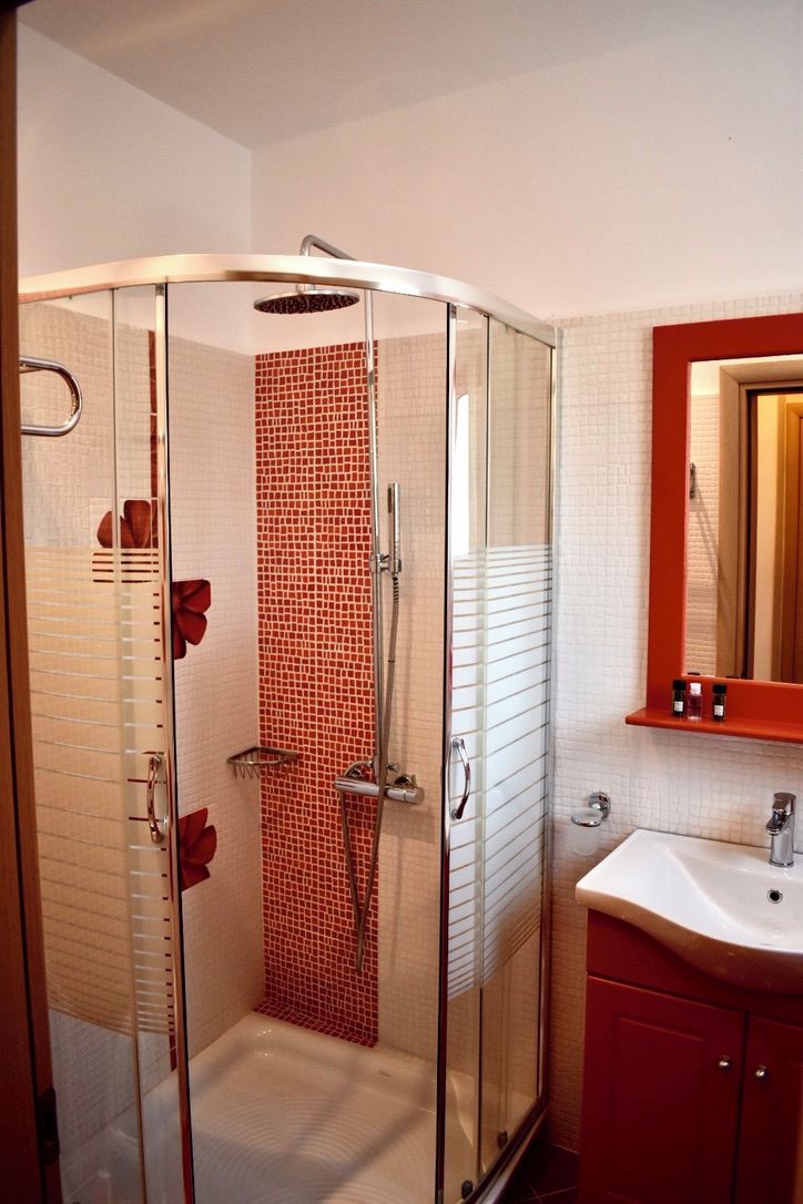 A glass-enclosed shower with a red-tiled wall, a white sink, and a mirror above it.