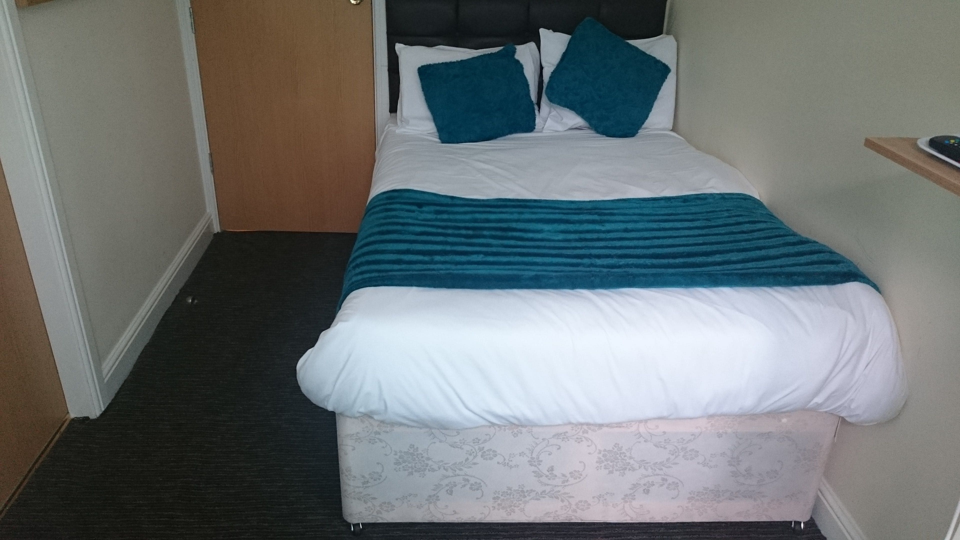 A neatly made bed with a teal blanket and two pillows, a patterned rug, and a wooden door in a hallway.
