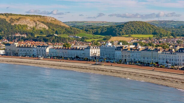 A coastal town with a row of white buildings along the beach, a clear view of the sea, and rolling hills in the background. - Elsinore Hotel (Llandudno)